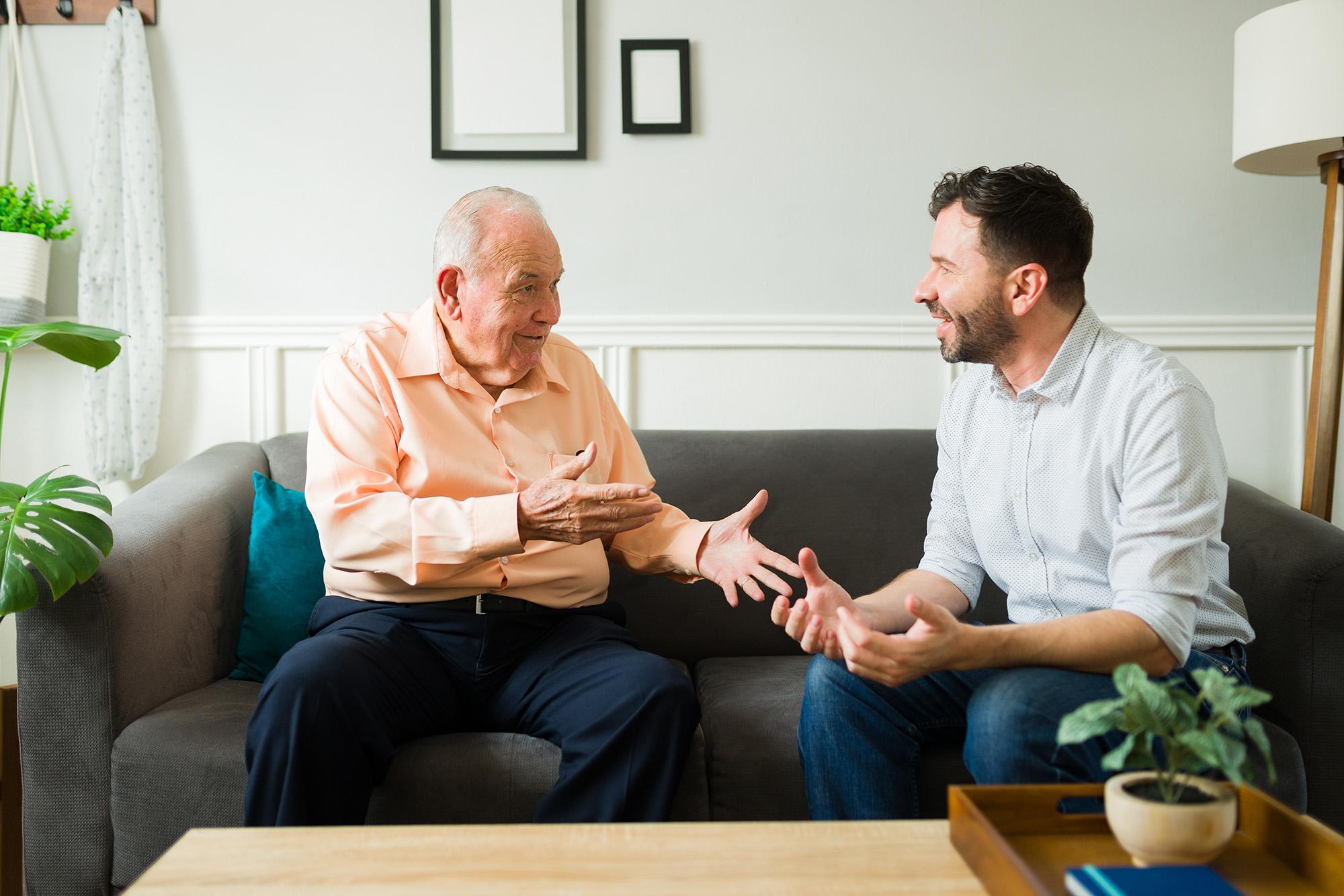 Two men sitting on a couch talking to each other.